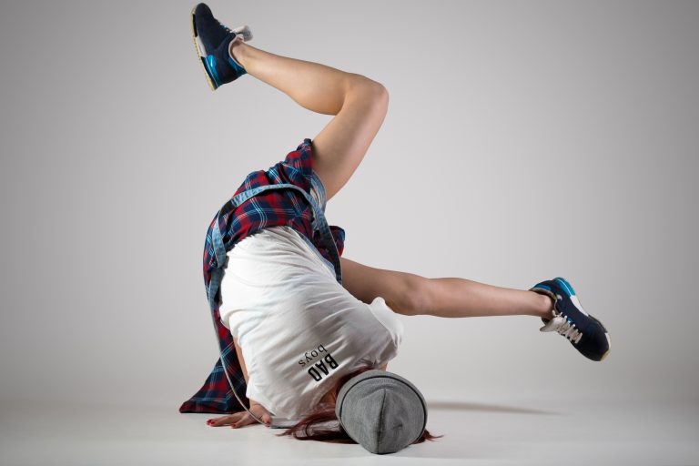 One fit young woman wearing casual plaid shirt performing breakdance moves on the floor. Modern style beautiful teen dancer working out, dancing. Full length image, studio gray background, back view